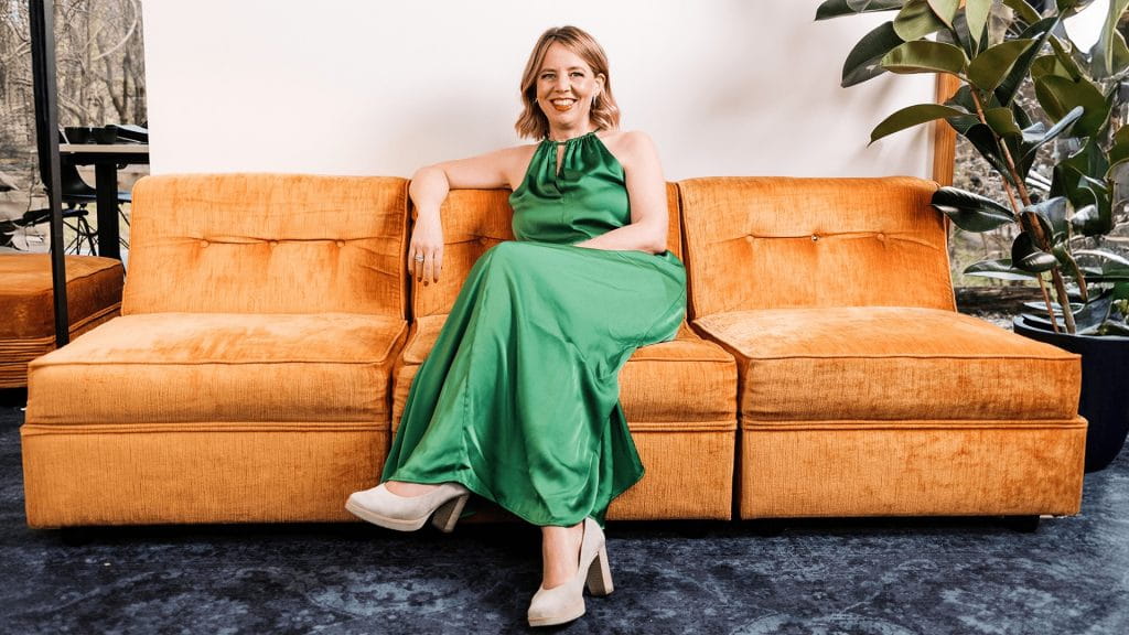 Mentorin sitzt in grünem Kleid auf einem senfgelben Sofa, entspannte Business-Porträtaufnahme in modernem Wohnzimmer-Ambiente